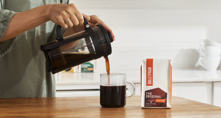 Une main versant une presse française de café dans une tasse à côté d'un sac de grains de café à l'épreuve des balles